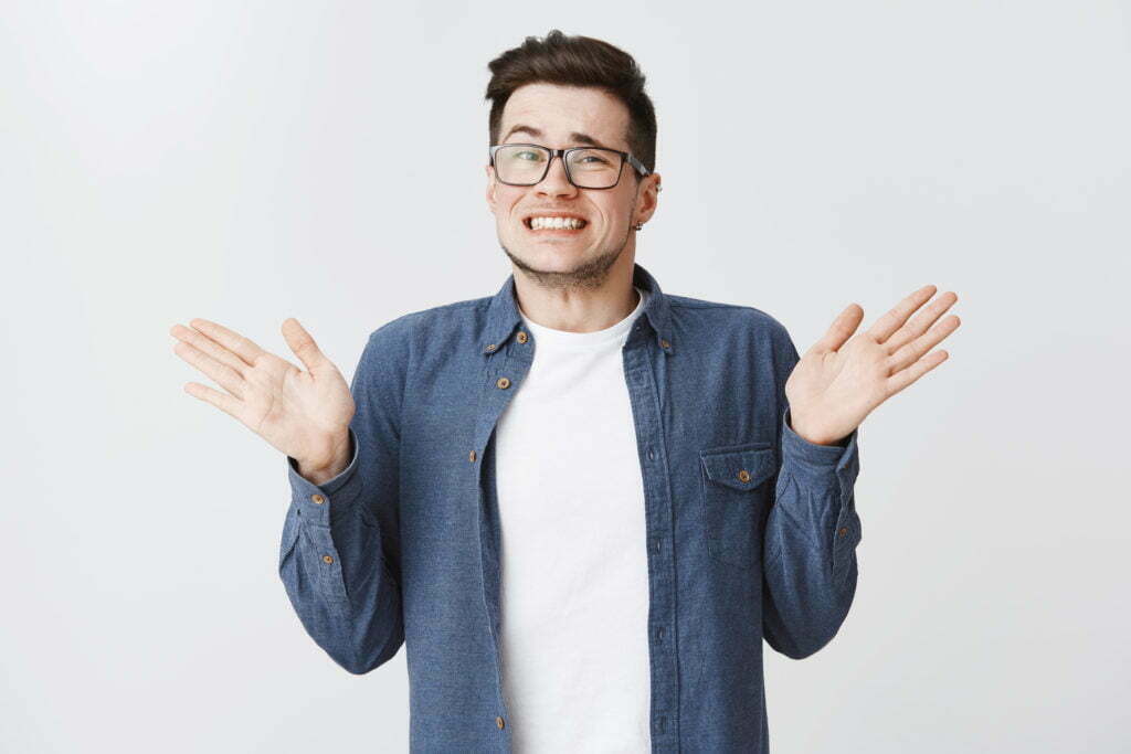 Sorry I messed up. Guilty young and handsome male in glasses and blue shirt raising hands near shoulders, shrugging making awkard smile as apologizing for making mistake over white background.