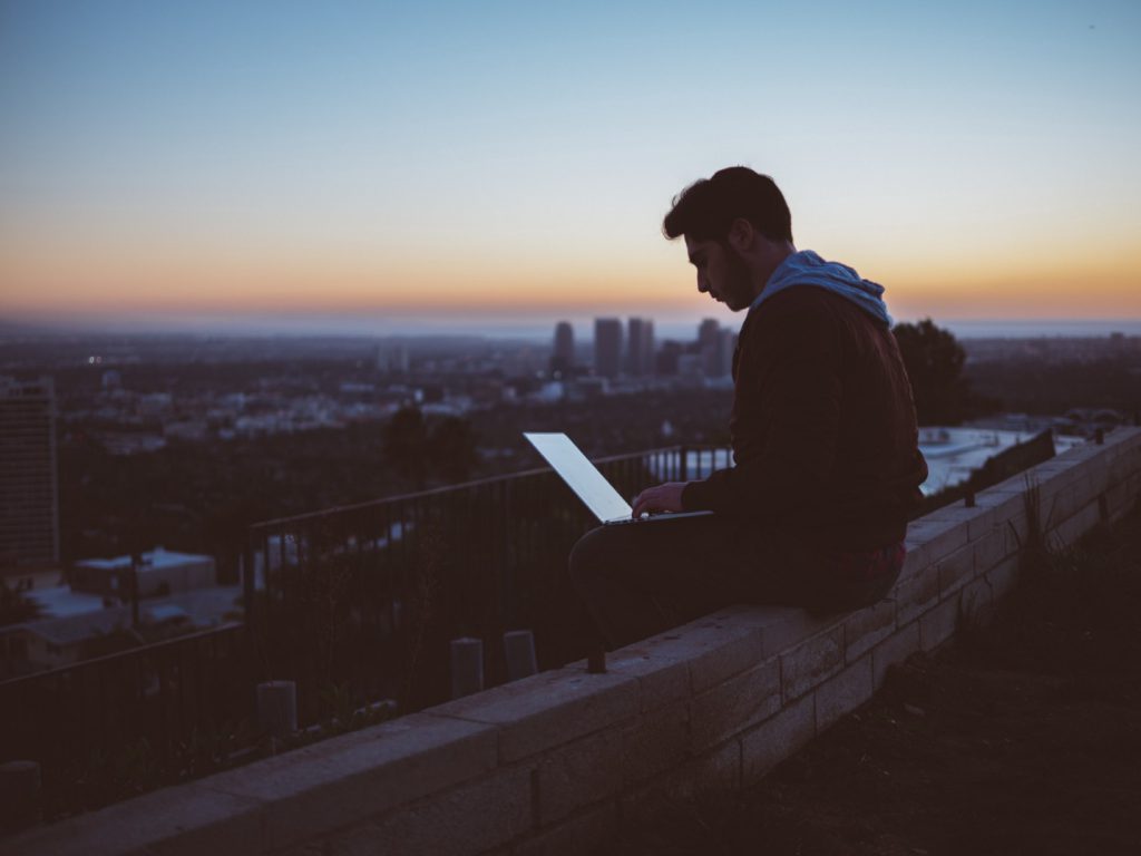 Young man sitting on the edge of a building with his computer on his lap