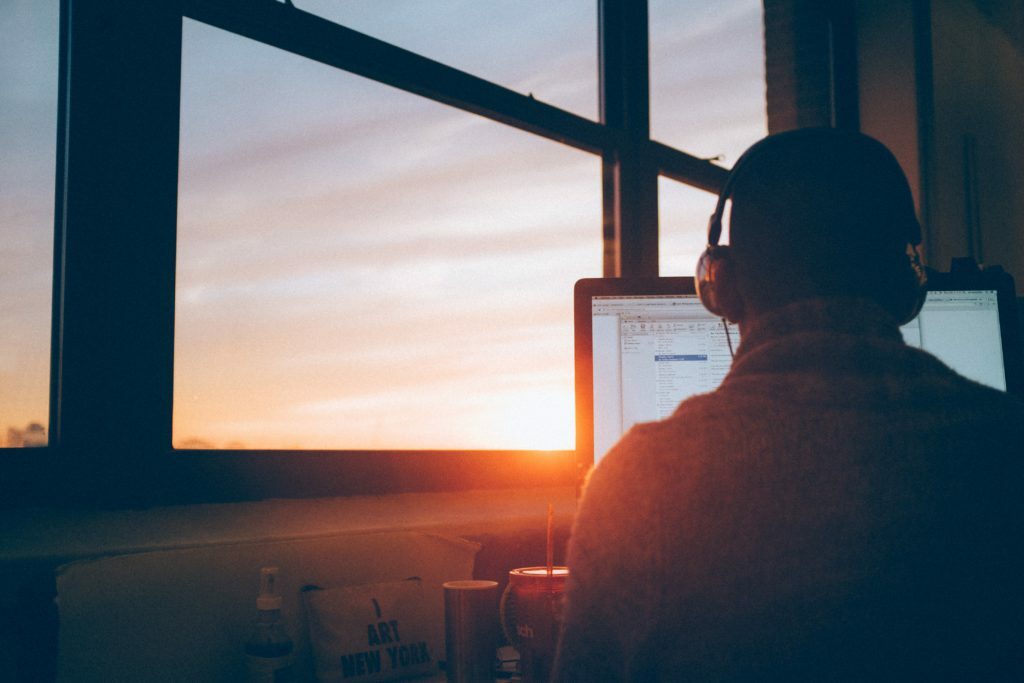 man working on his computer with his headphones in, near a window at sunset
