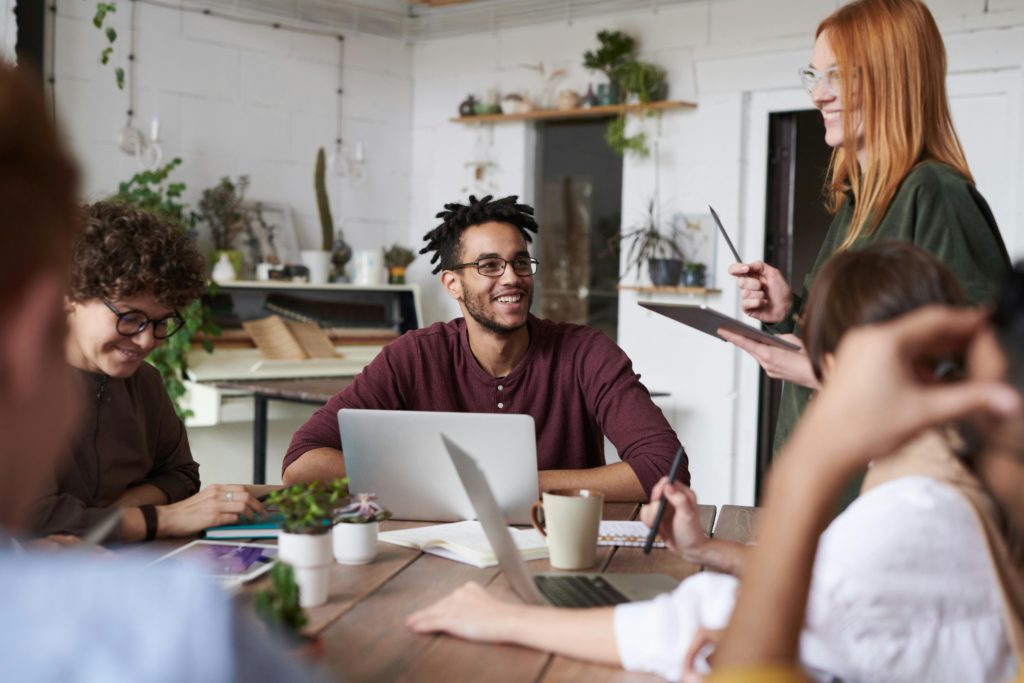 A diverse group of colleagues engaging in a positive discussion around a table, with laptops, notepads, and plants visible. One person is smiling, while another is standing and holding a tablet, facilitating the conversation in a casual office setting with a bright, relaxed atmosphere